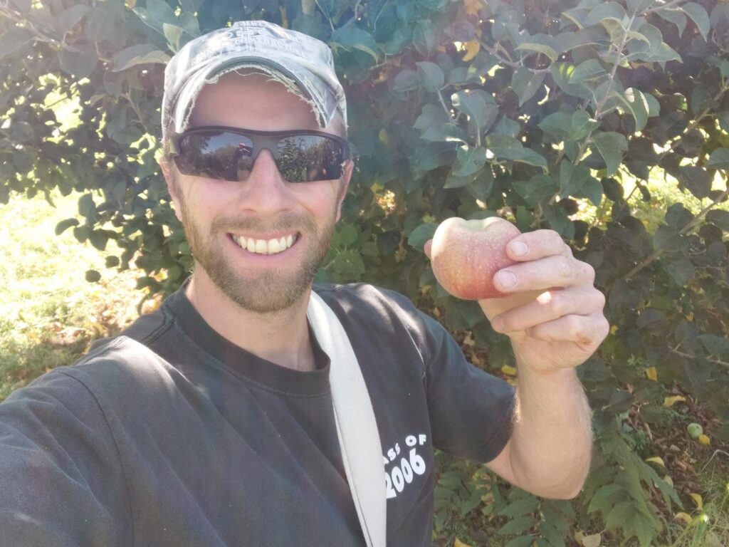 A man wearing a baseball cap and glasses is holding an apple picked at Linden Hall Farm and smiling into the camera.