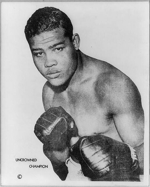 A black and white image of boxer Joe Louis wearing boxing gloves.