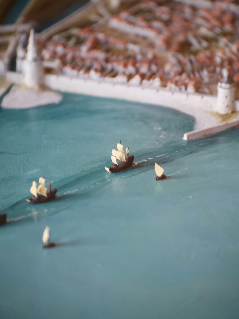 A scene of miniature sail boats approaching a coastal city.