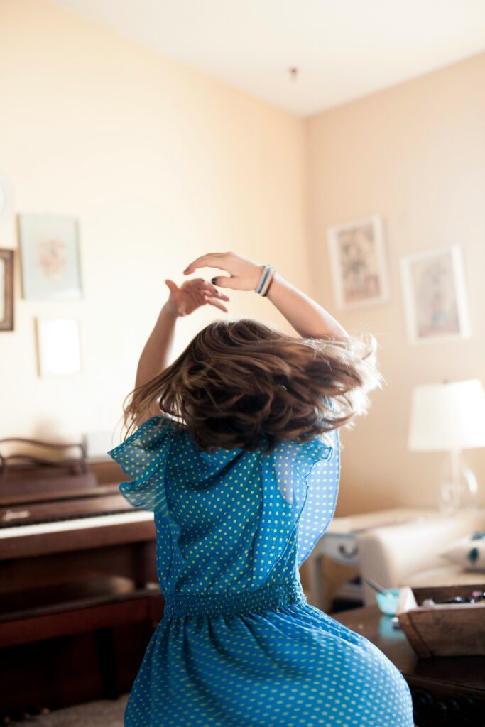 A happy girl in motion, she is seen twirling in a brightly lit room and is not facing the camera.